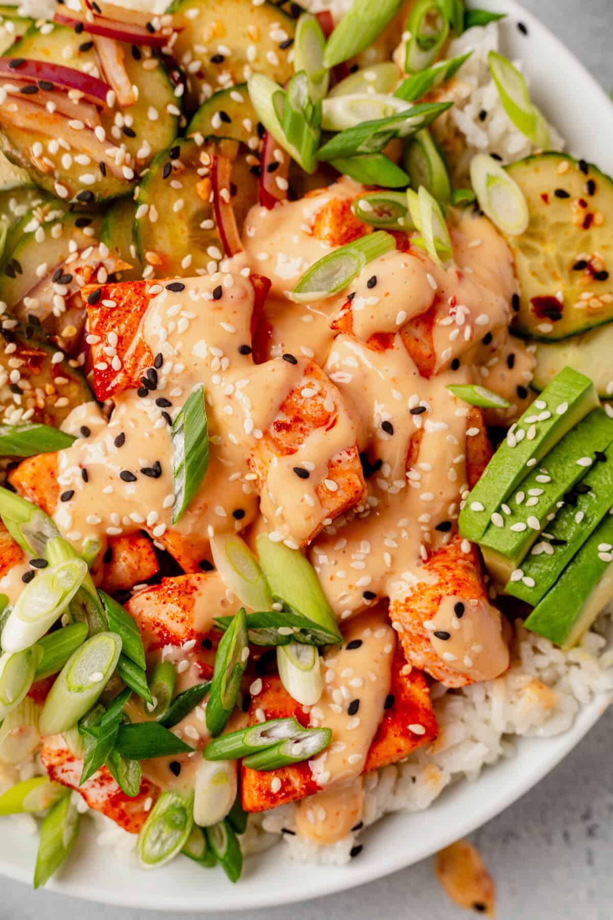 Close-up of a bang bang salmon rice bowl, featuring glazed salmon bites topped with creamy spicy sauce, sliced green onions, sesame seeds, avocado slices, and cucumber salad with red onion, all served over white rice.