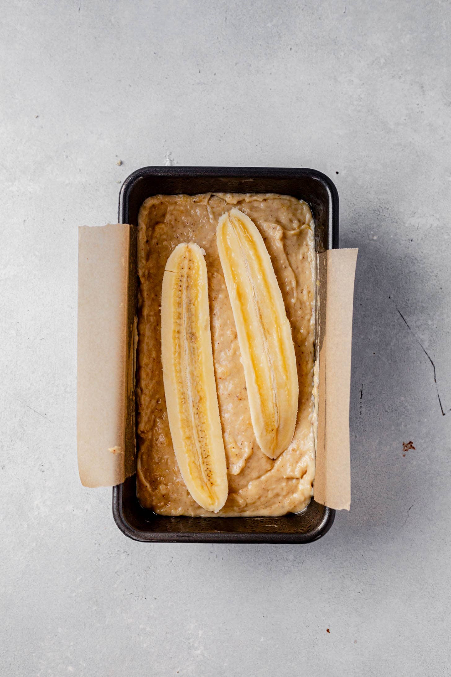 Banana bread batter in a parchment-lined loaf pan topped with two halved bananas lengthwise, ready to go into the oven.