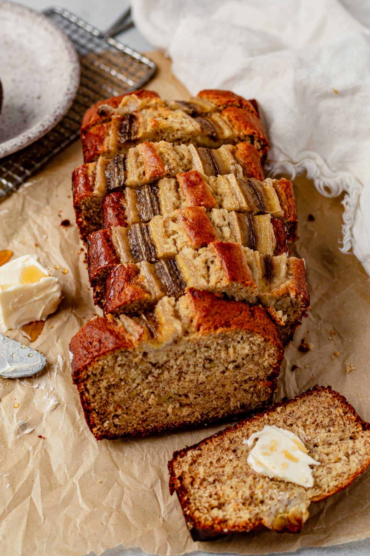 Freshly sliced loaf of brown butter banana bread topped with caramelized banana pieces, served on parchment paper with a buttered slice in front and a pat of butter nearby.