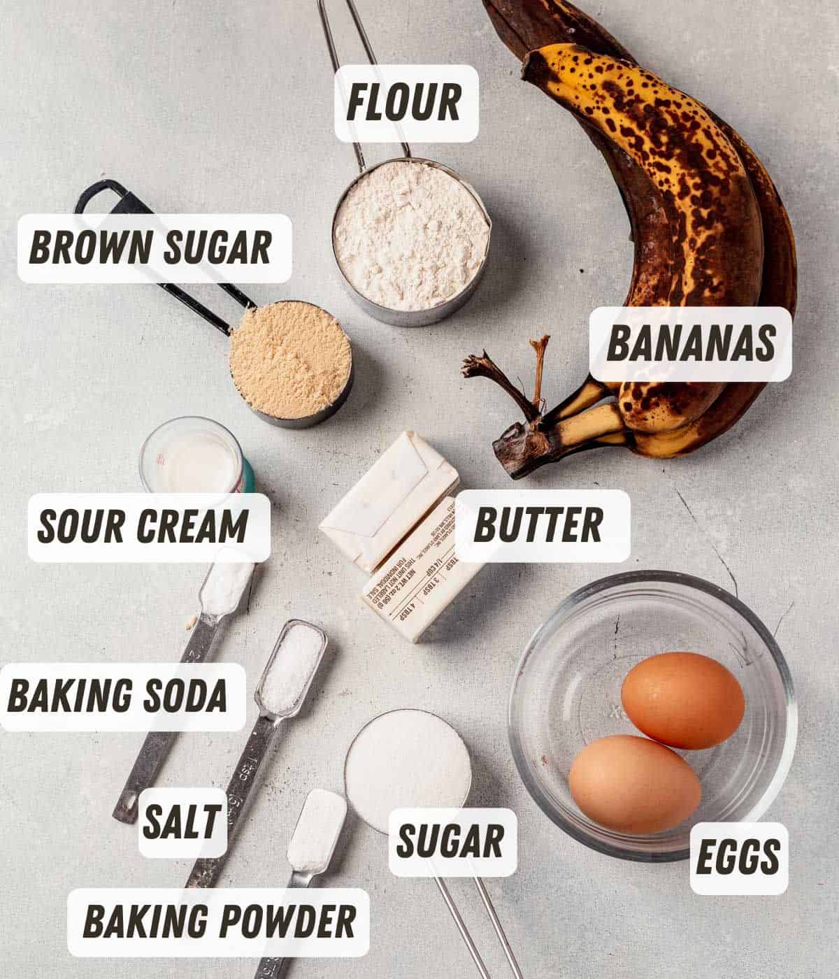 Flat lay of labeled banana bread ingredients including ripe bananas, flour, brown sugar, sour cream, butter, baking soda, salt, sugar, baking powder, and eggs on a light gray background.