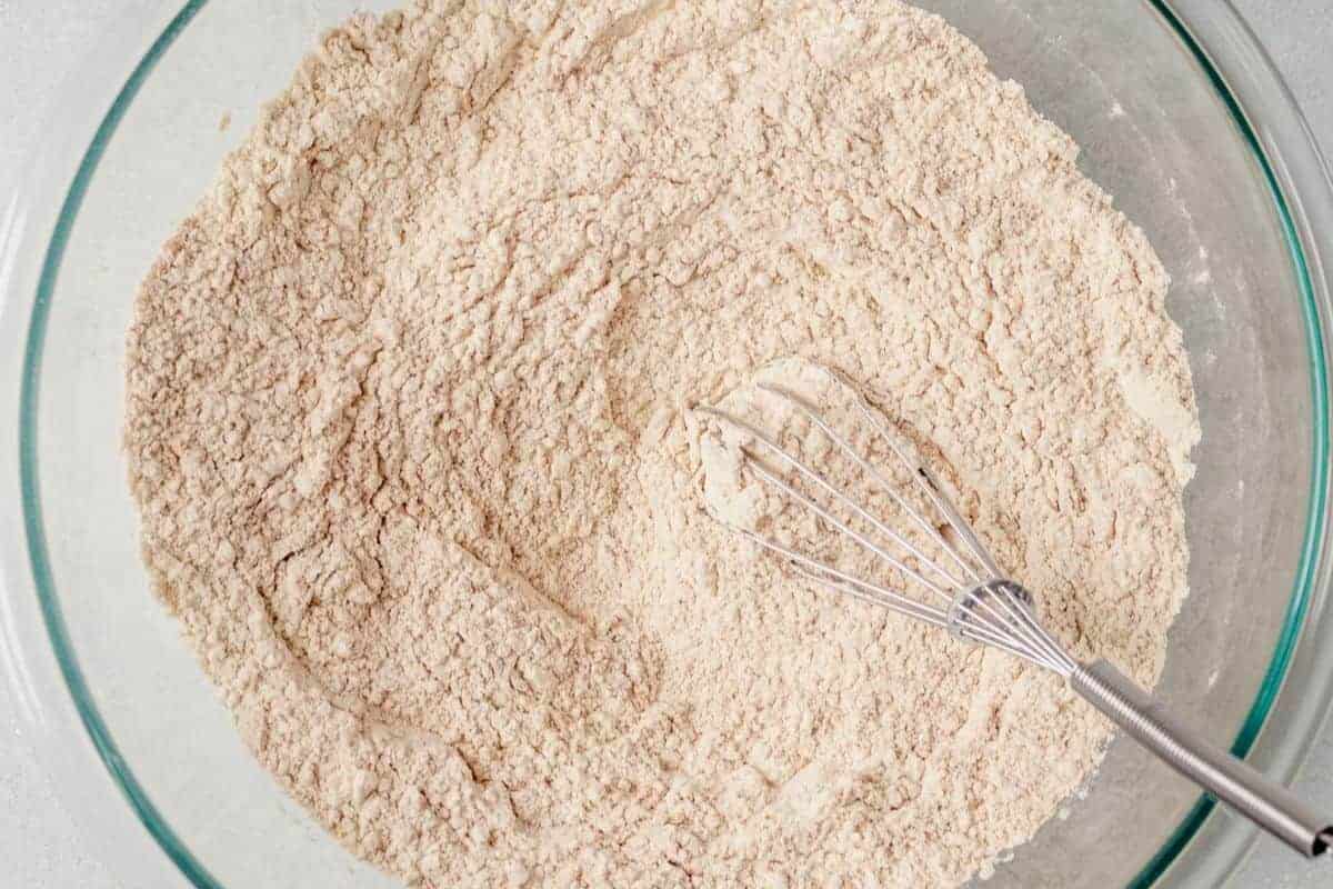 Glass mixing bowl filled with whisked dry ingredients, including flour, baking powder, and spices, with a metal whisk resting inside.