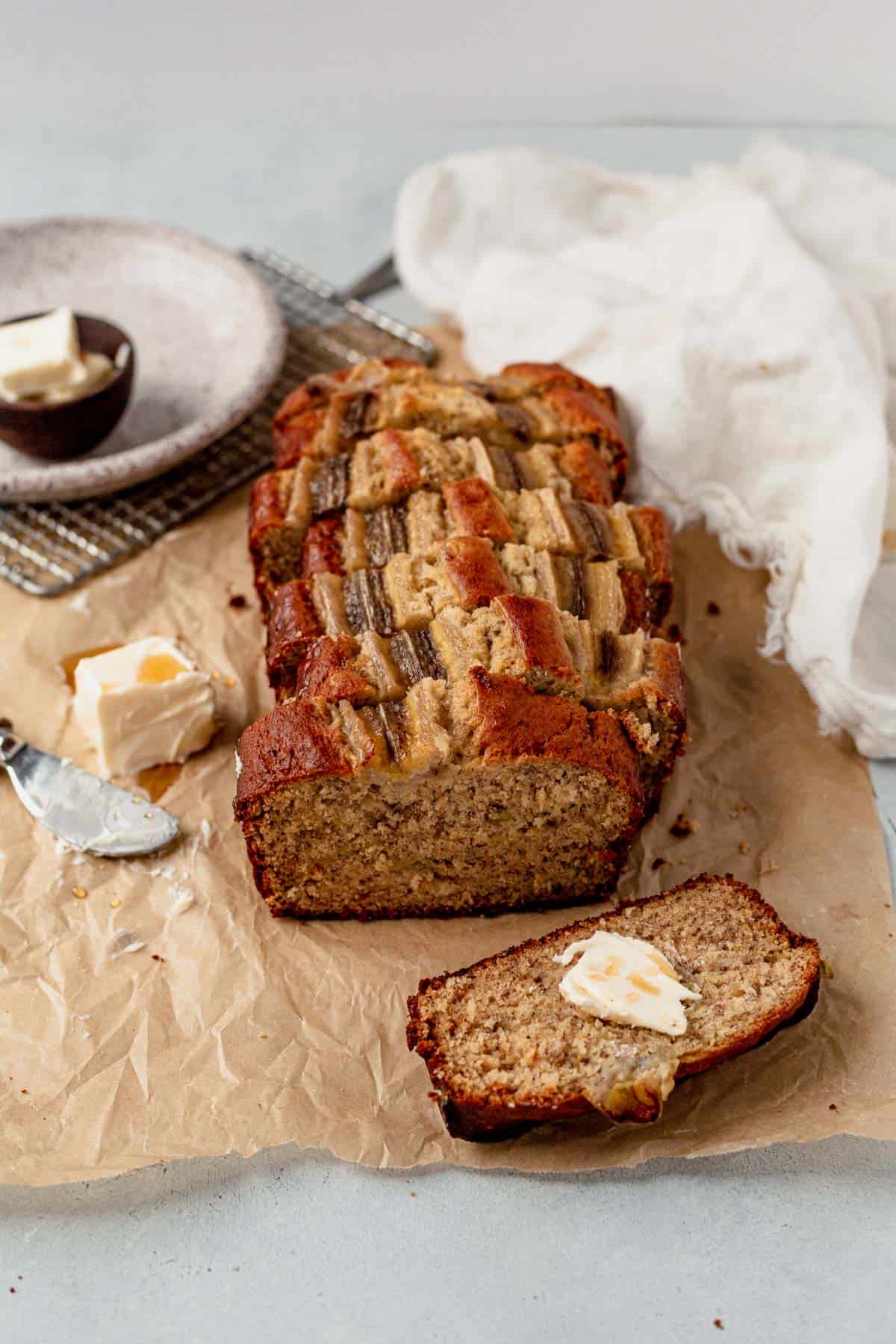 Freshly baked loaf of brown butter banana bread on parchment paper with a sliced piece topped with melting butter and honey, served beside a dish of butter.
