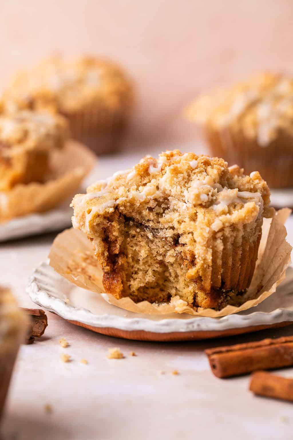 Close-up of a coffee cake muffin with a bite taken out, revealing a moist cinnamon-sugar swirl inside. The muffin is topped with crumbly streusel and drizzled icing, sitting in a peeled-back liner on a stacked ceramic plate. Cinnamon sticks and muffin crumbs are scattered around for a warm, inviting scene.