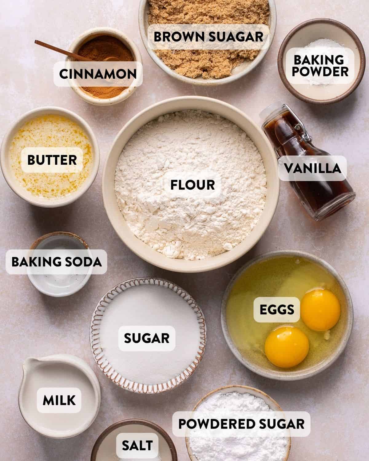 Flat lay of common baking ingredients arranged on a light countertop, including a large bowl of flour at the center, surrounded by brown sugar, white sugar, powdered sugar, cinnamon, melted butter, two cracked eggs in a bowl, milk, vanilla extract in a small glass bottle, baking powder, baking soda, and salt. Each ingredient is labeled clearly in bold text.