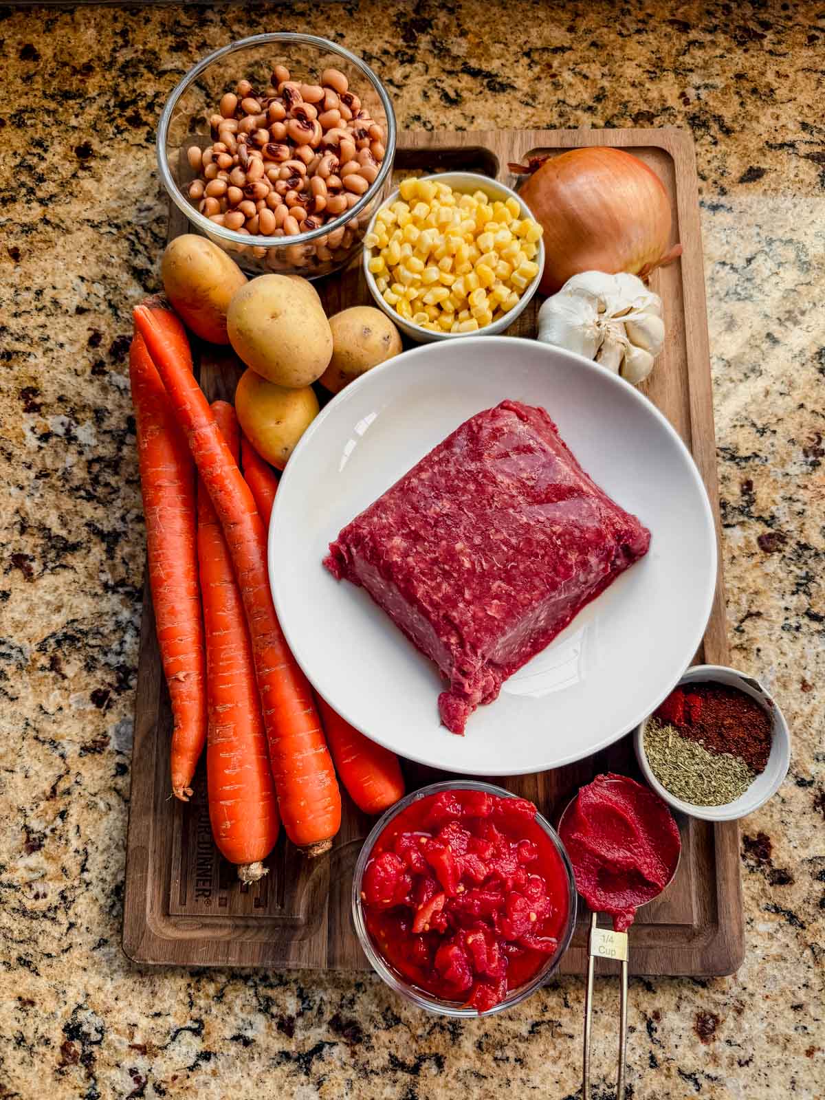 ground beef, carrots, corn, tomatoes potatoes, onion, and garlic on a cutting board.