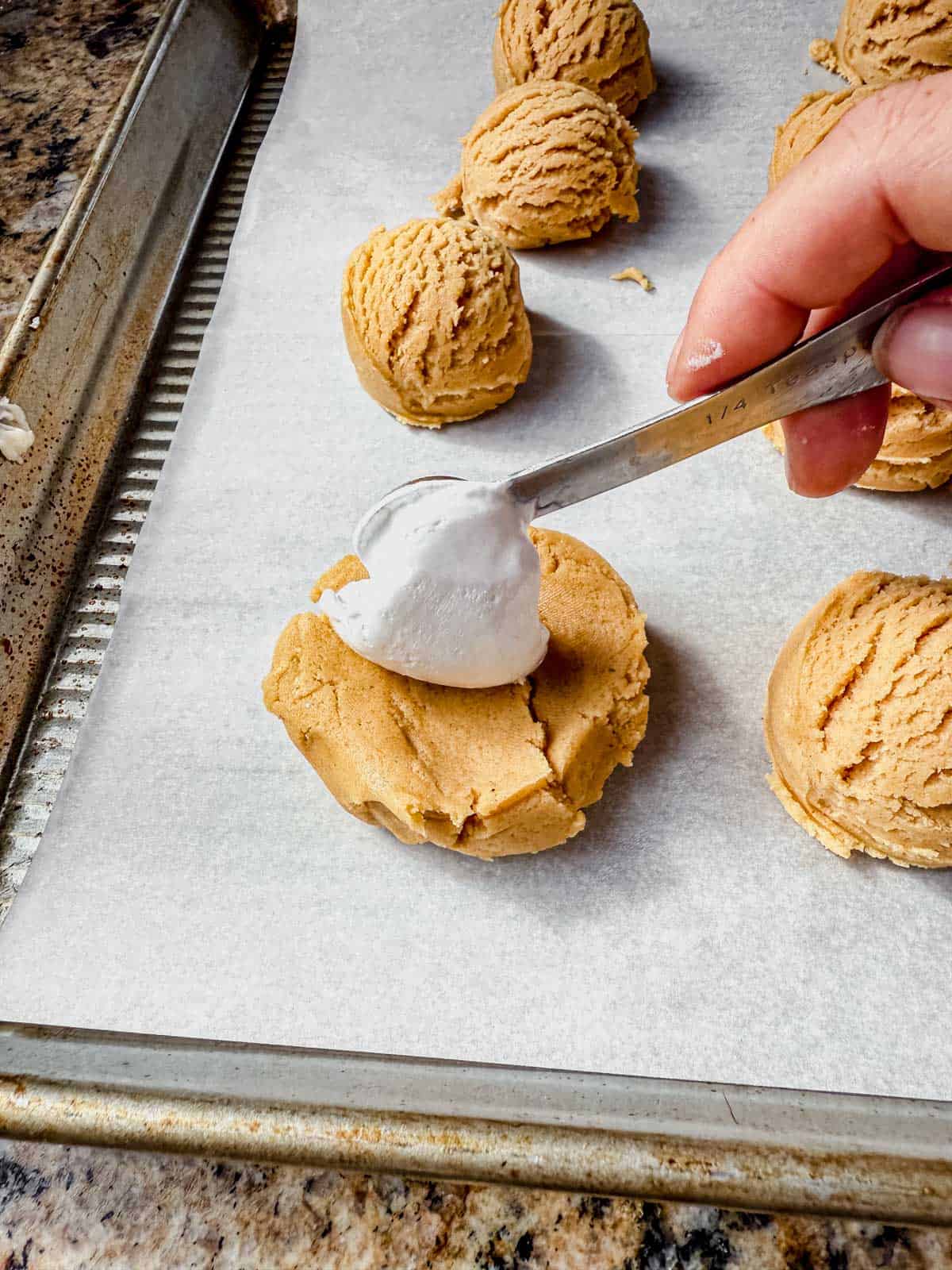 back of a small spoon laying marshmallow fluff on the center of fluffernutter dough balls on a parchment lined baking sheet.