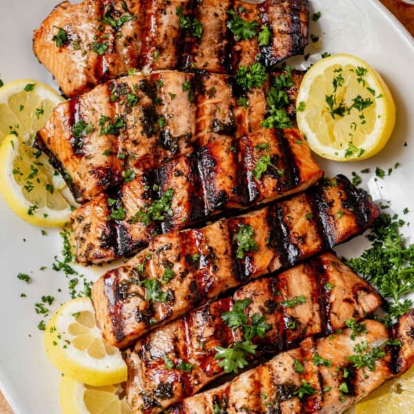 Grilled salmon filets with charred grill marks, garnished with fresh parsley and lemon slices on a white serving platter.