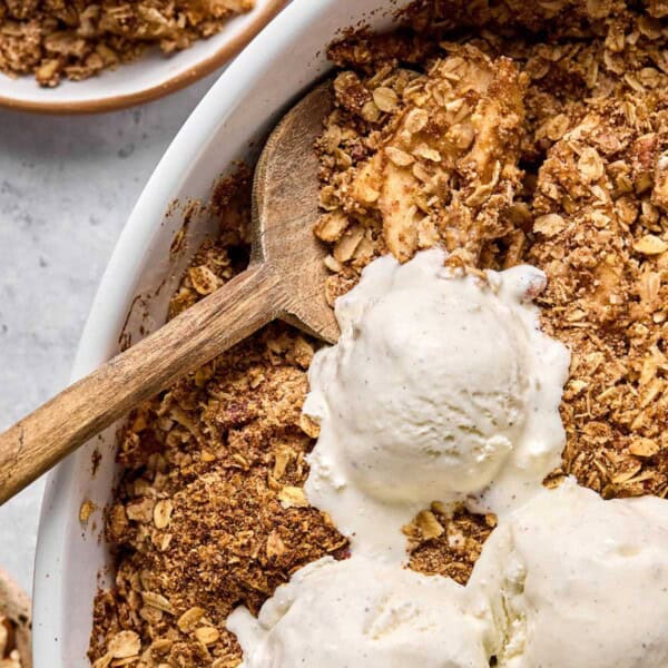 baked healthy apple crisp in a baking dish with three scoops of vanilla ice cream on top and a wooden spoon digging in