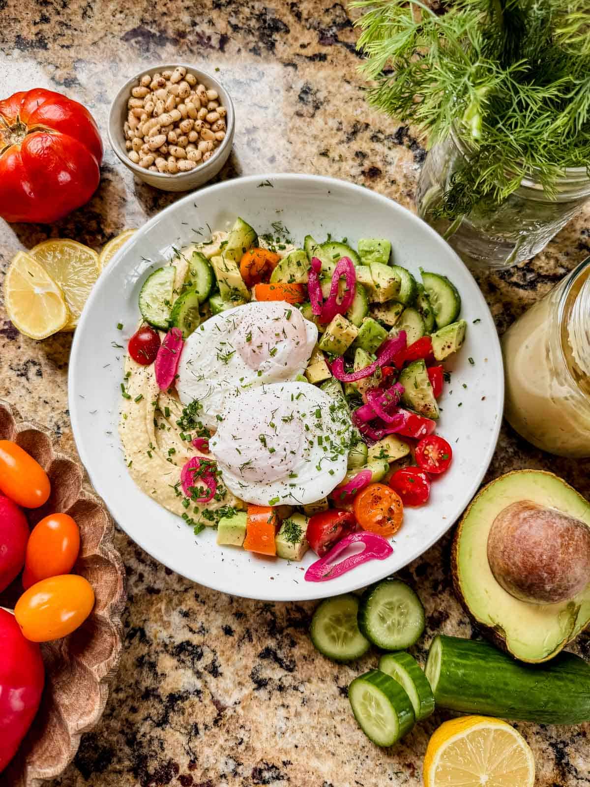a mediterranean breakfast bowl on the counter with fresh herbs, pine nuts, and avocado around it.