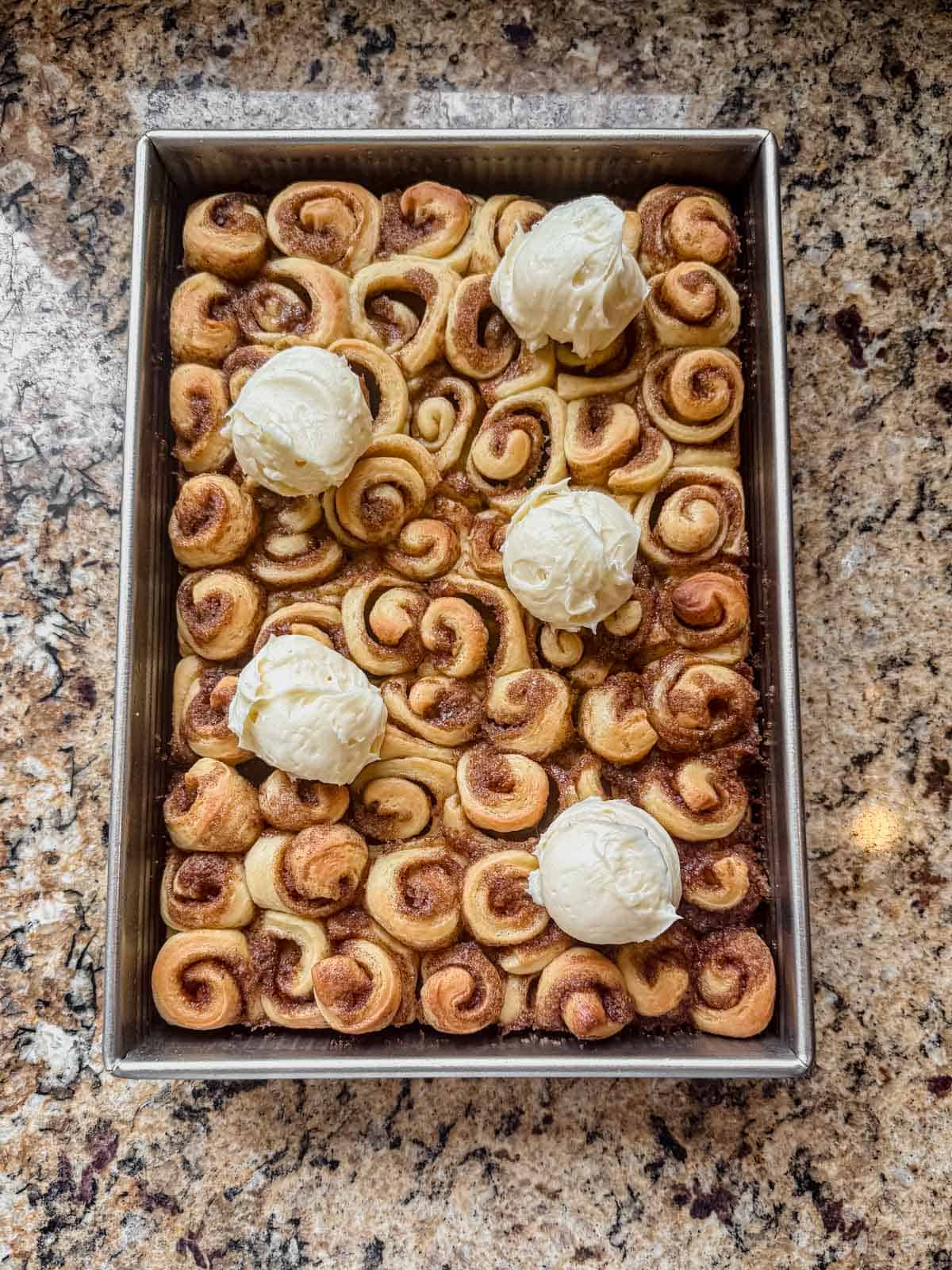 warm mini cinnamon rolls in a 9x13 pan with scoops of cream cheese frosting on top.