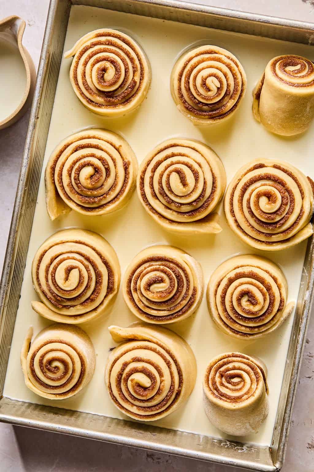 unbaked cinnamon rolls in a baking dish with heavy cream all around them.