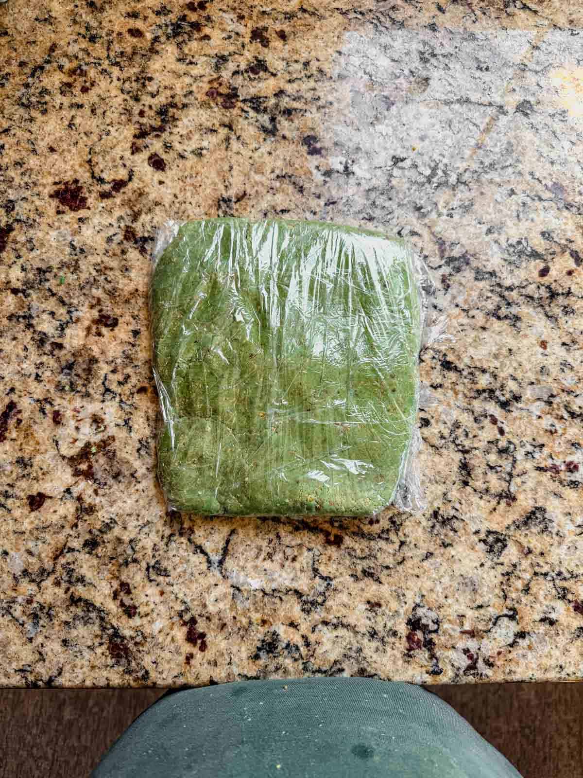 pistachio linzer dough flattened into a rectangle inside of plastic wrap on a granite countertop, prepared to chill