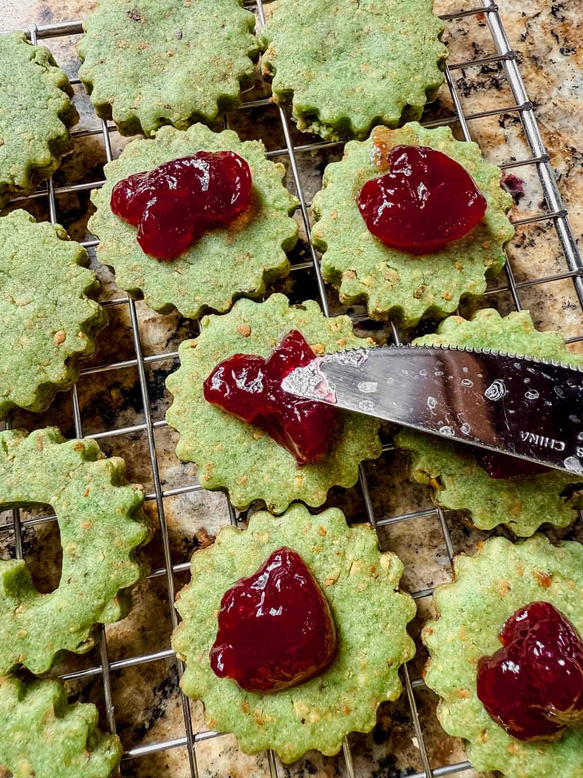 butter knife spreading raspberry jam in a heart shape onto baked pistachio linzer cookies